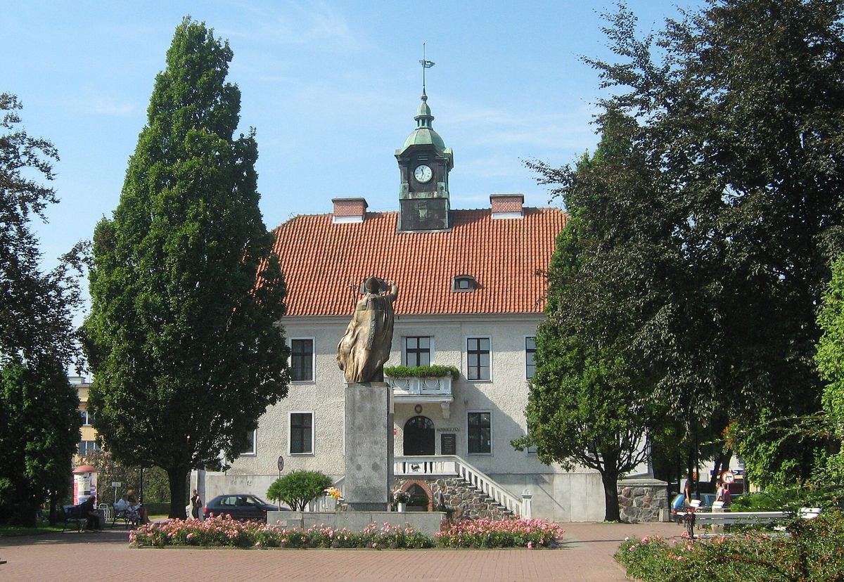 Weißes Rathaus mit Uhrturm und rotem Dach, davor ein Denkmal und Bäume