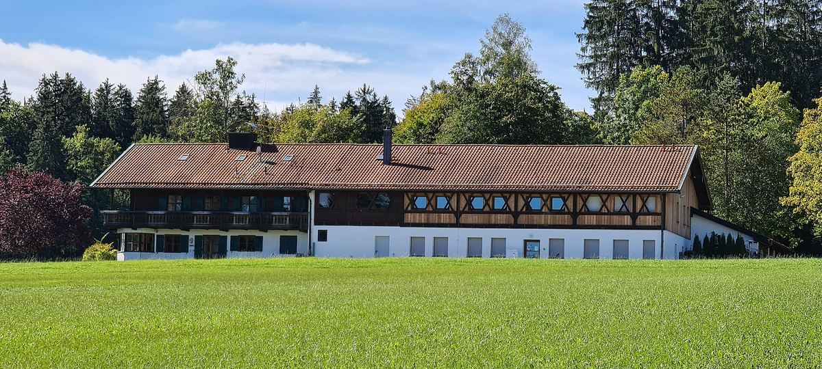 Langgestrecktes Landhaus mit Holzbalkon hinter einer Wiese, Bäume im Hintergrund